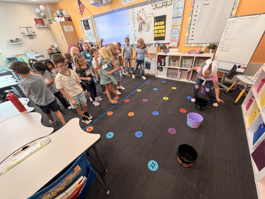 A classroom with children standing in a circle around a floor with colorful dots. One child is picking up objects from a bucket.