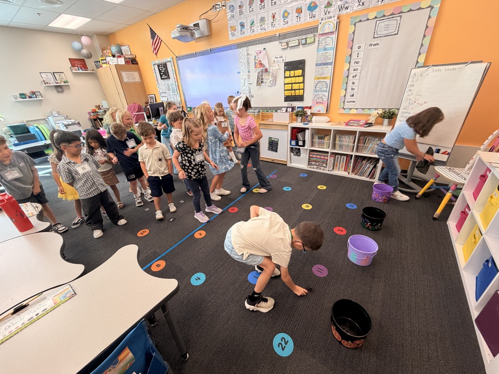 Several children in a classroom playing a game with buckets and colored discs on the floor.
