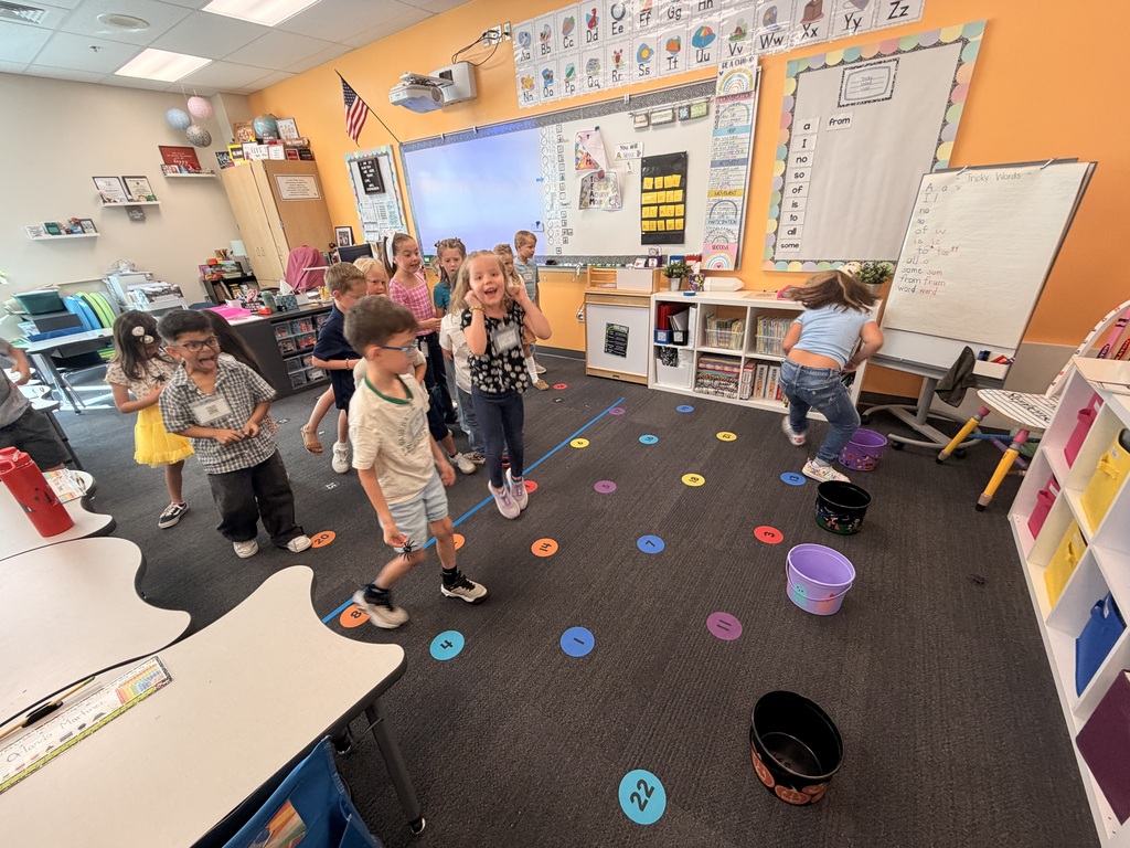 Children playing on a floor with multi-colored circles and buckets. A classroom with white boards, desks, chairs, and a flag.