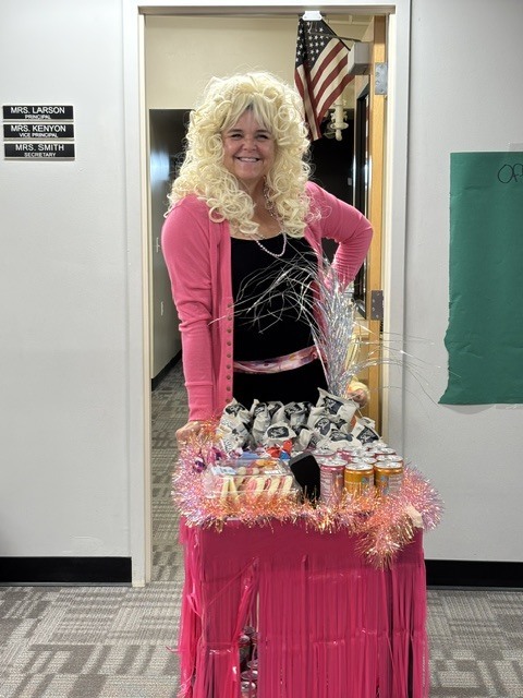A person with blonde hair stands at a pink table with various items. Behind them is an open doorway with an American flag.