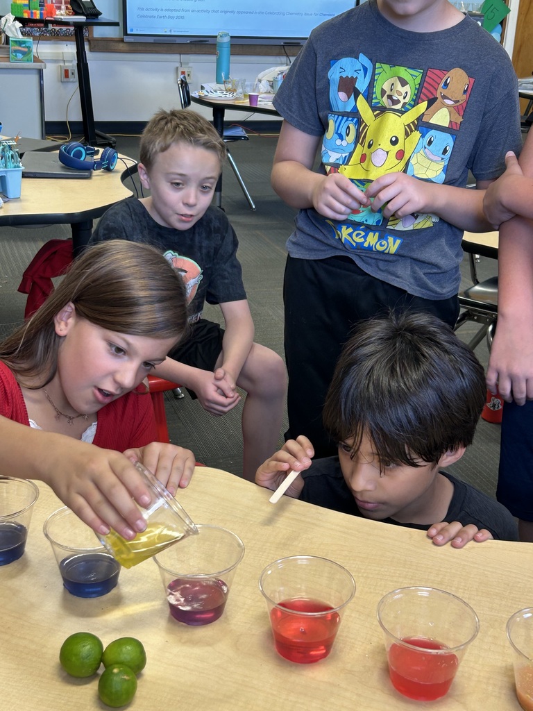 Four kids are gathered around a table with cups of colored liquids. One girl pours liquid from a jug.