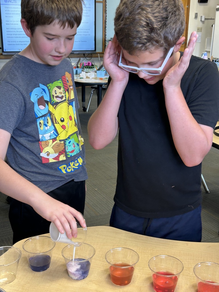Two boys, one wearing glasses, pour colored liquids into cups on a table.