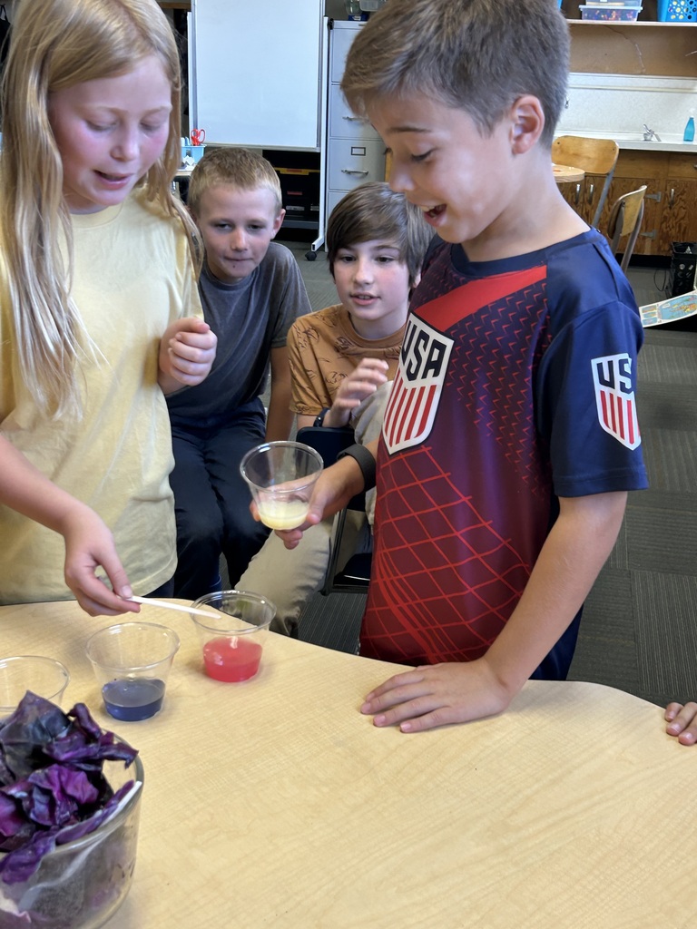 A group of children in a classroom, one holds a small cup with red liquid, another has a blue liquid.