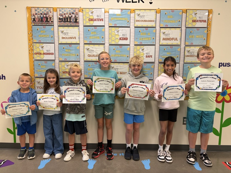 Seven children, some in shorts, stand in a row, holding certificates. Behind them is a wall with colorful boards and text.