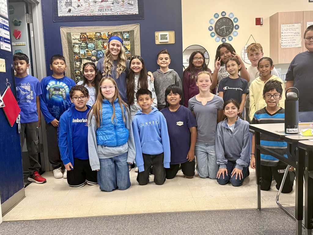 A group of children in a classroom, smiling and posing for a photo. One adult stands behind them.