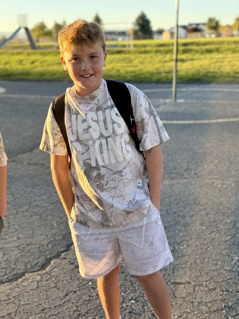 A boy smiles while standing in a parking lot. He wears a "Jesus is King" shirt and shorts.