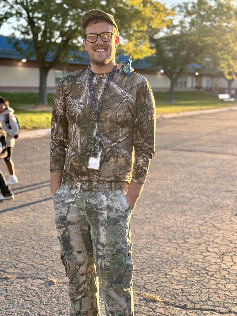 A man with a smile stands in a parking lot wearing camouflage clothing. Trees and a building are in the background.