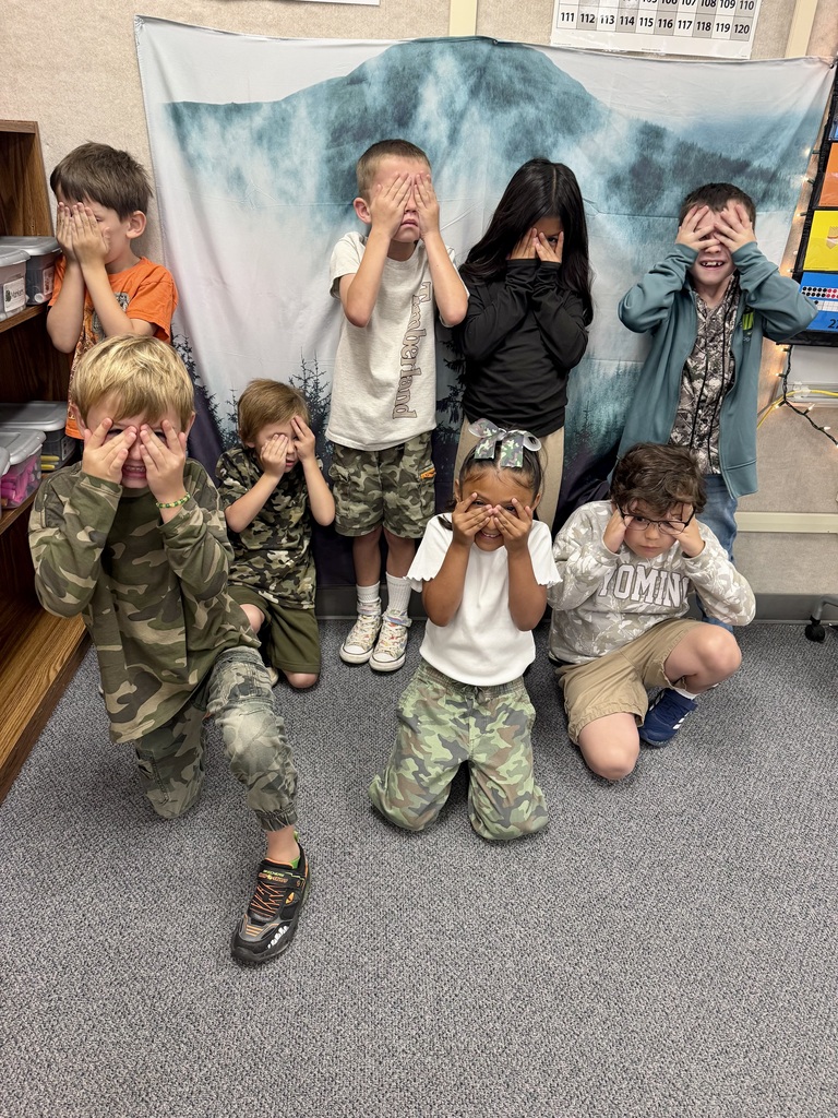 Nine children in camouflage and other clothing cover their eyes, kneeling or squatting. A white backdrop is behind them.