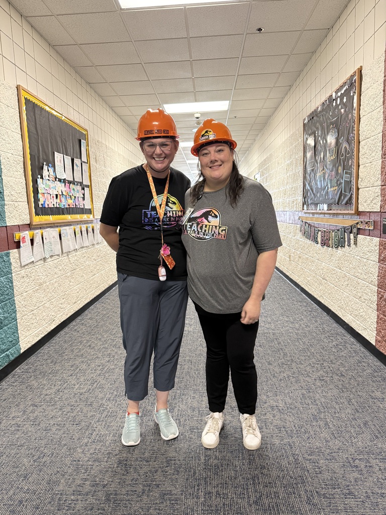 Two women, one in orange and the other in gray, stand in a hallway. Behind them, a bulletin board and various pictures adorn the walls.