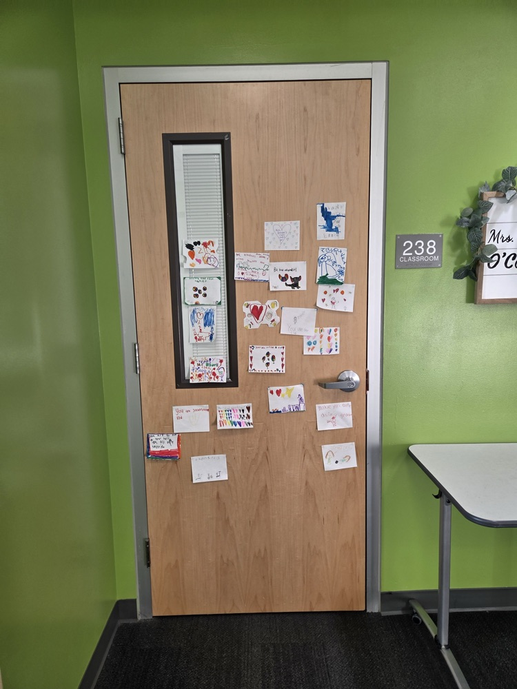 A wooden door with a glass window. Many colorful stickers cover the door. Green wall and black floor. A small white table is to the right.