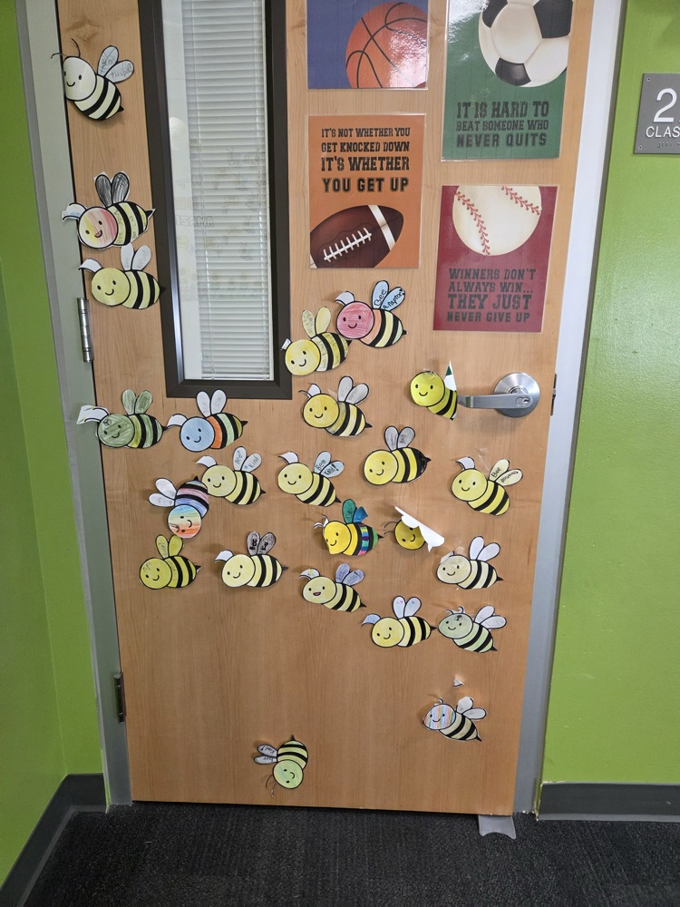 A wooden classroom door with green walls. It is decorated with many stickers of bees and motivational quotes.