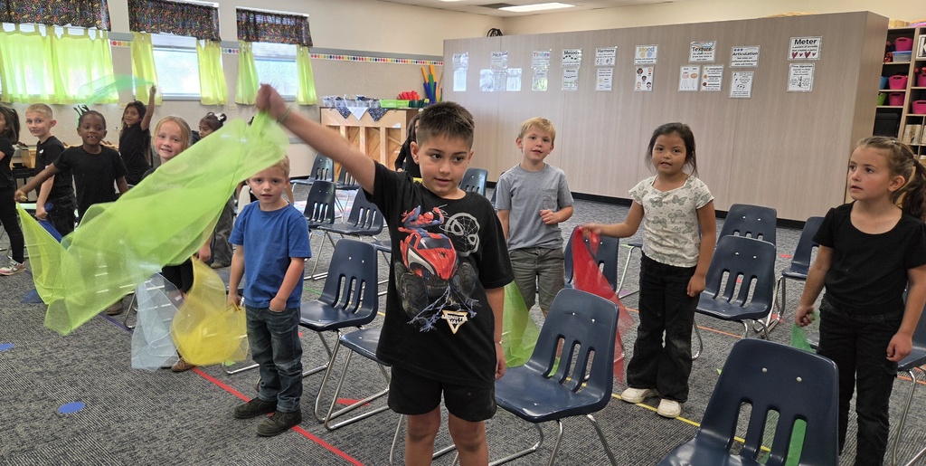 A classroom with children in black and blue shirts. One child holds a green ribbon while others stand around chairs.