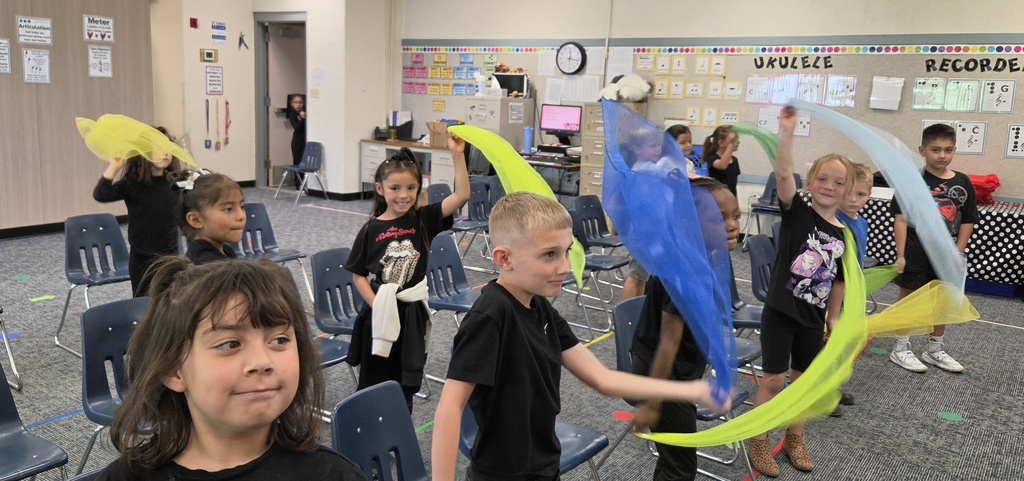 A group of children in black t-shirts are practicing dance moves in a classroom with blue chairs.