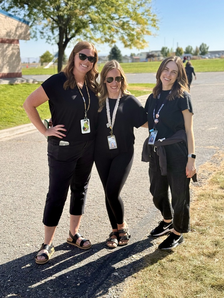 Three women stand together in an outdoor setting, wearing matching black clothing and sunglasses. They are in a paved area with grass and trees in the background.