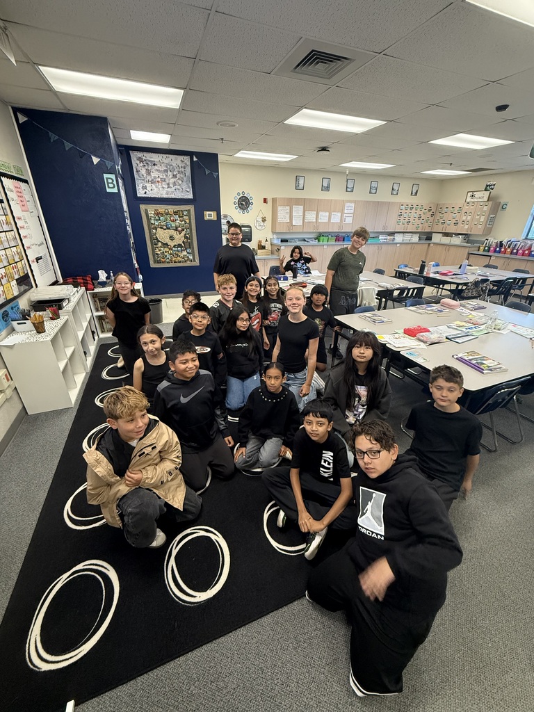Students in black shirts sit or stand on a rug in a classroom with tables and shelves.