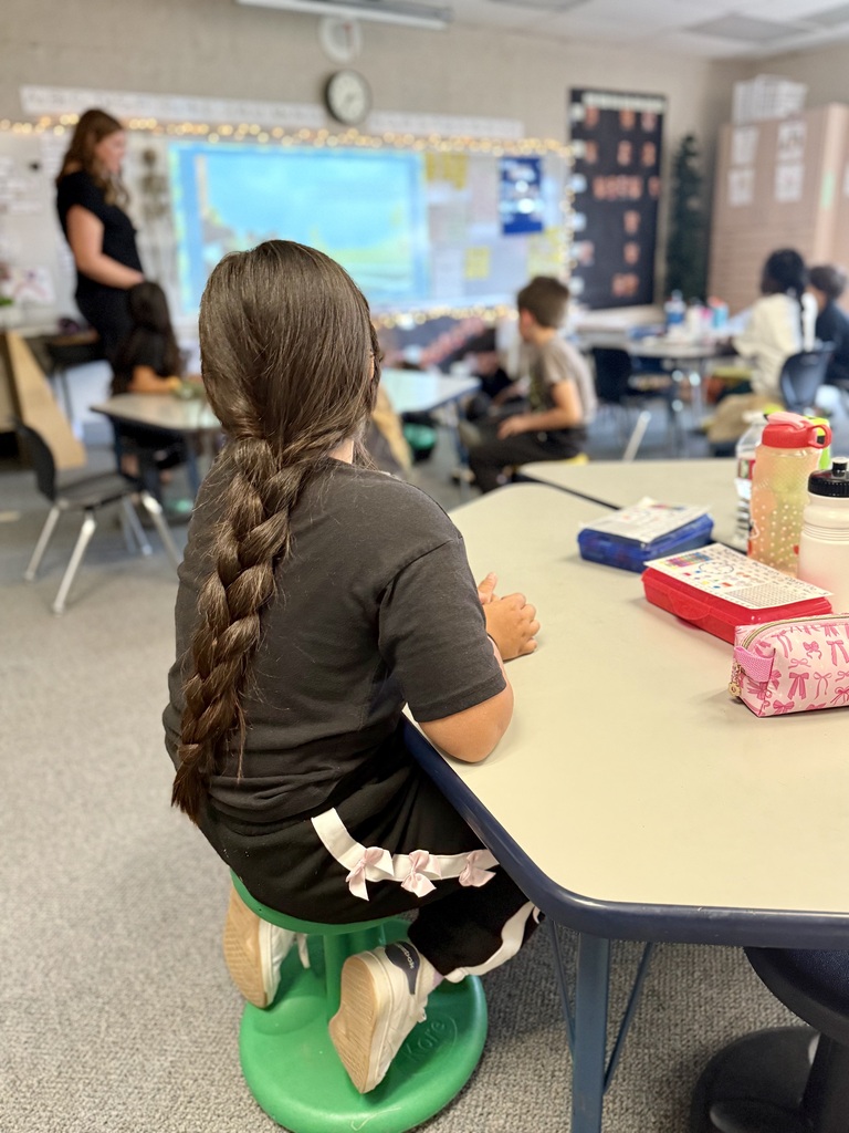 A child with long braided hair seated on a stool at a table in a classroom.
