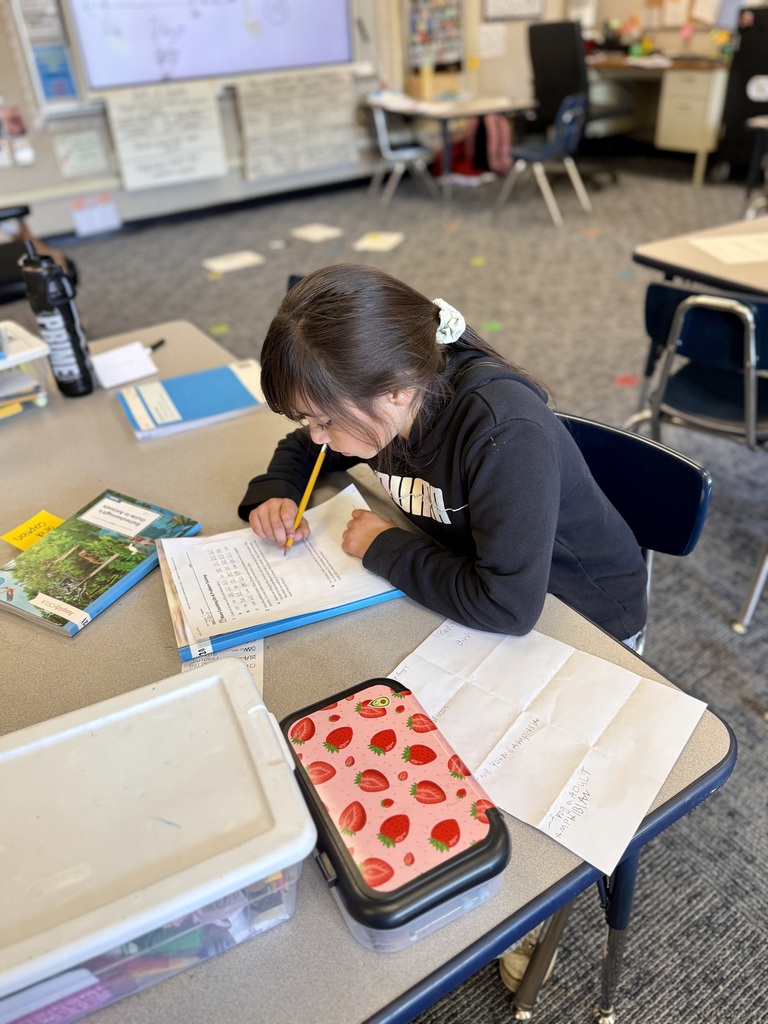 A student sits at a desk in a classroom, writing with a pencil on a piece of paper.