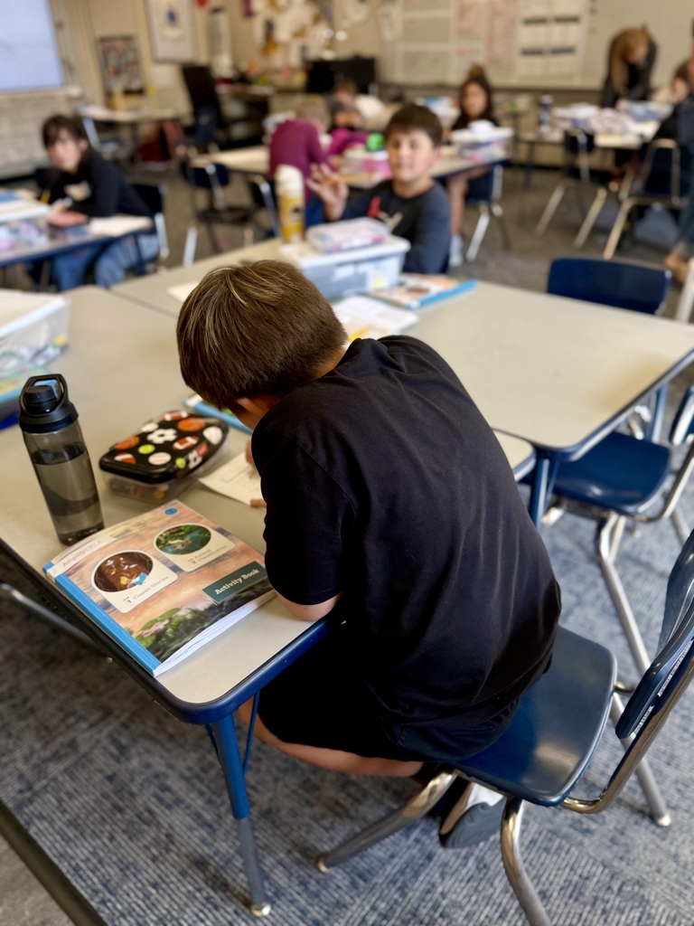 A student seated at a desk in a classroom, focused on reading a book. Other students sit behind him.