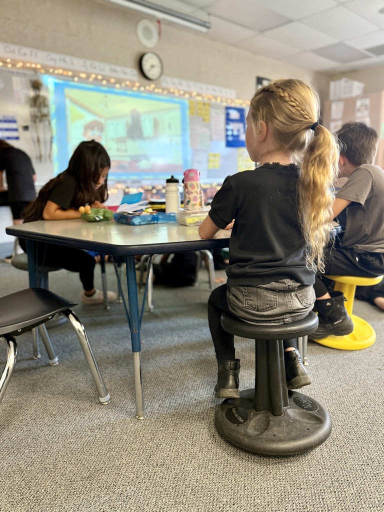 Three children sit at a table with a projector screen in the background. Two are seated on stools.