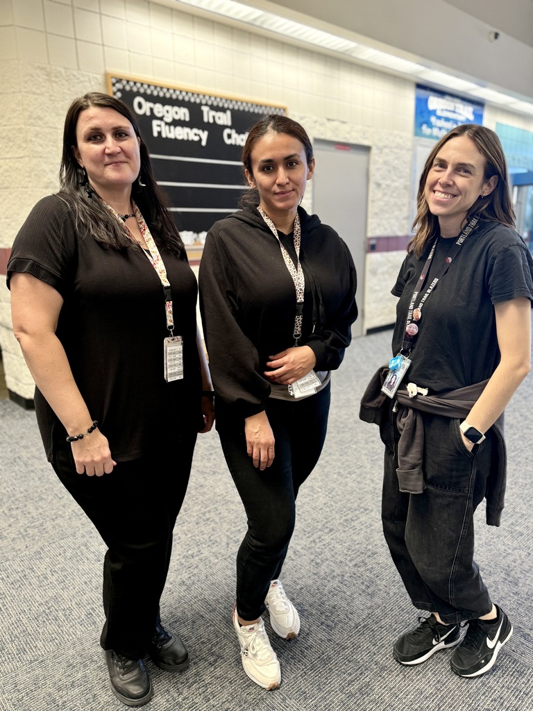 Three women, all dressed in black, stand next to each other. They wear ID cards and shoes. Behind them is a sign that reads "Oregon Trail Fluency Chat."