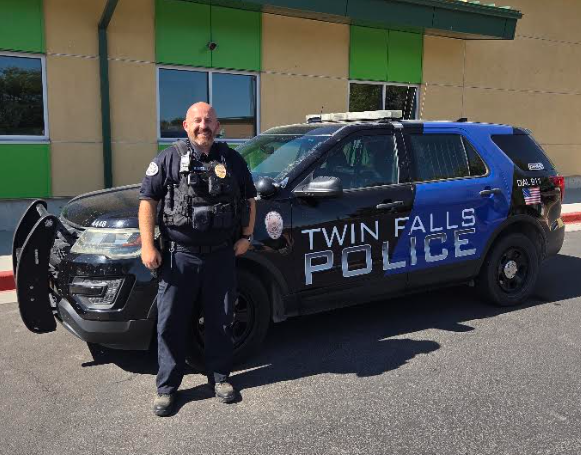 A male police officer standing in front of a black and blue police car