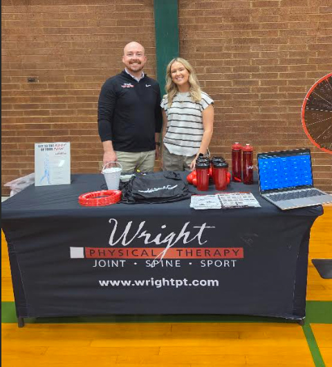 a picture of a male and female standing behind a table with a black table cloth in a school gym