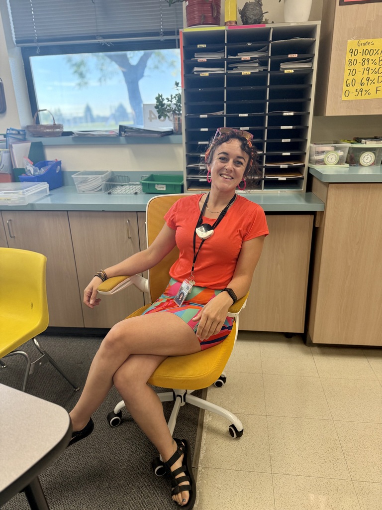 Woman in bright orange shirt sits in a yellow chair in a classroom with yellow chairs.