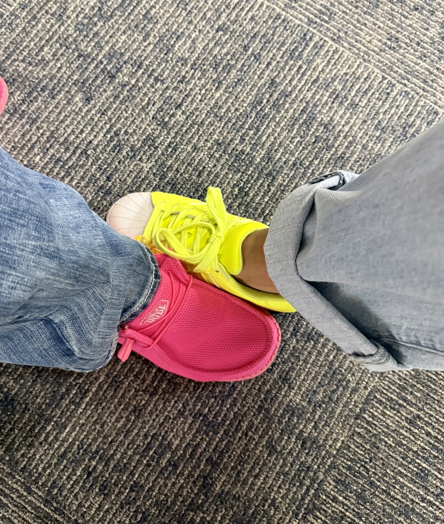 Two individuals standing close together. One is in yellow shoes, the other in pink. Both are wearing jeans. Background is carpeted floor.