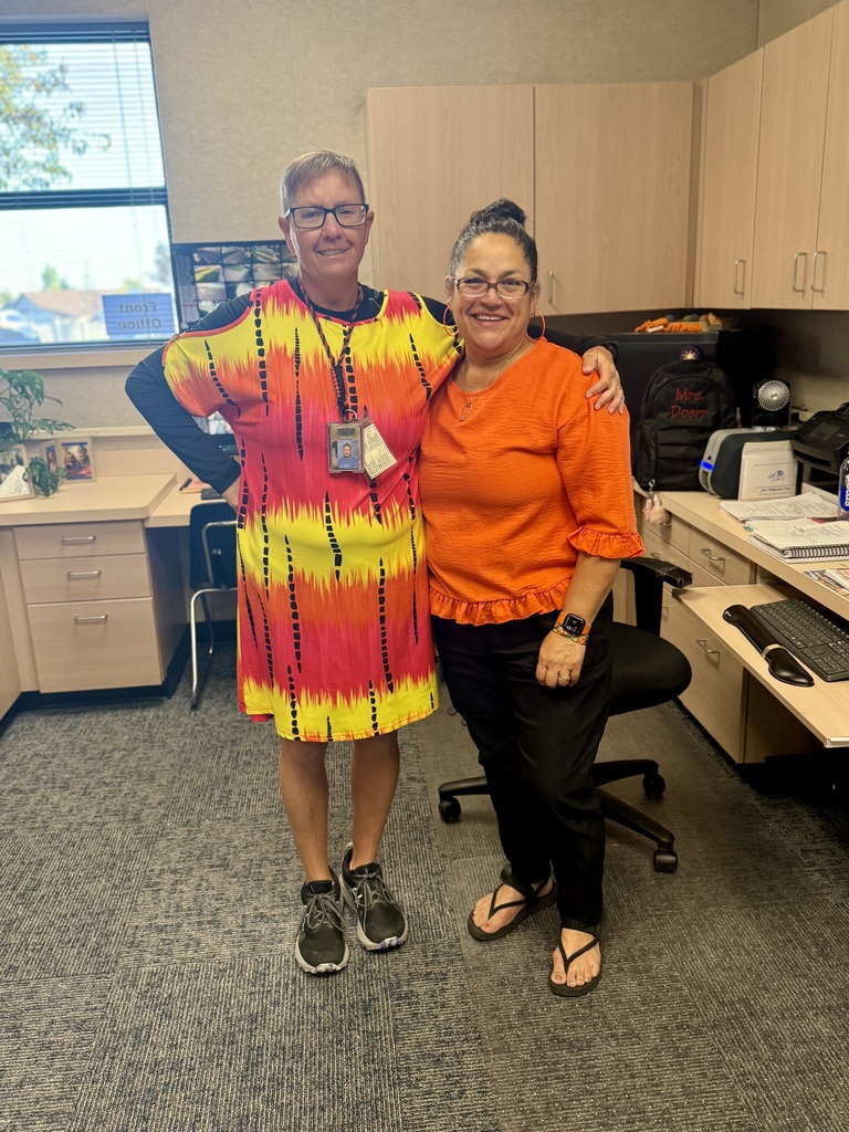 Two women standing in an office with a window. One wears a yellow and orange dress, the other an orange top. Desks, chairs, and cabinets are behind them.