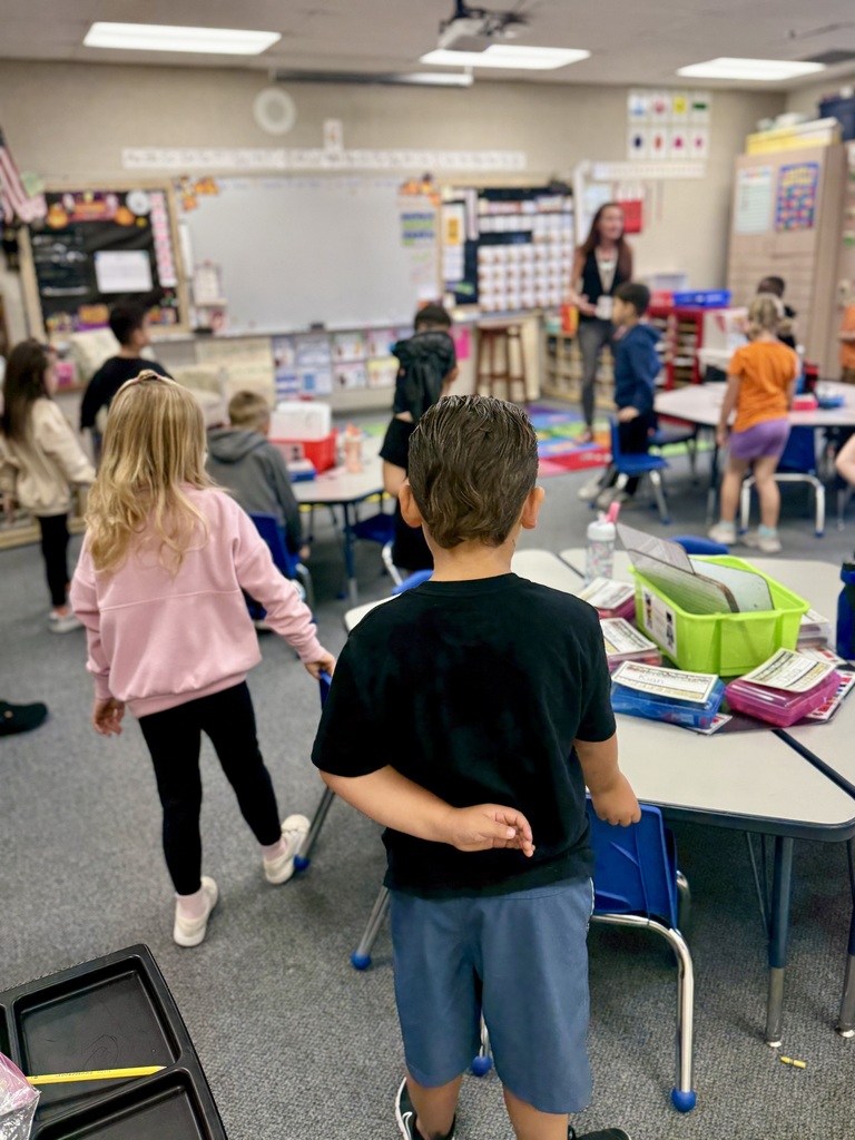 Children in a classroom with desks and a teacher. Bright lights and a whiteboard are visible.