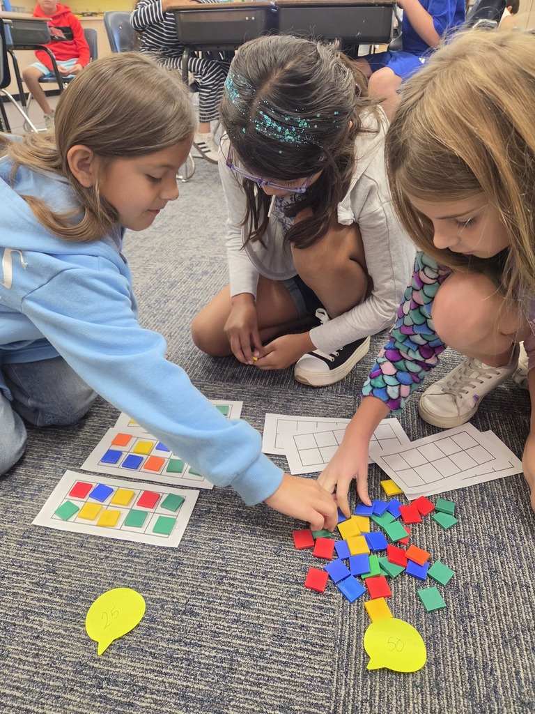 Three young girls in a classroom setting, using colorful geometric shapes on a floor mat.