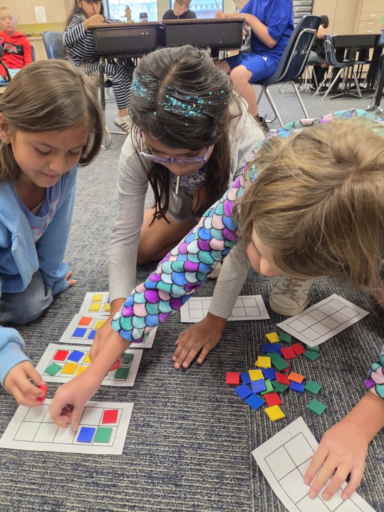 Three girls, one wearing glasses, work on a puzzle with colorful squares on the floor.