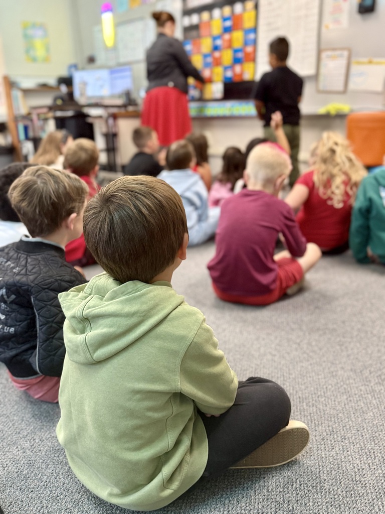A group of children sits on the floor in a classroom. A teacher stands behind a computer.