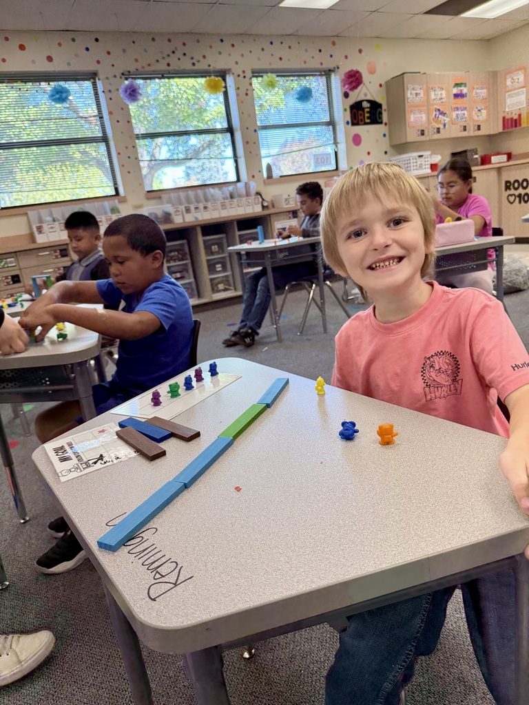 Children play with colorful blocks and pieces on a table in a classroom. Some sit at desks, others stand.