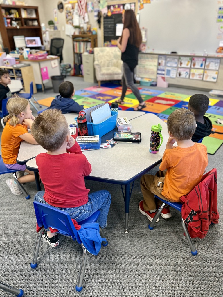 A classroom with students sitting at tables. A woman is walking towards the front of the room.