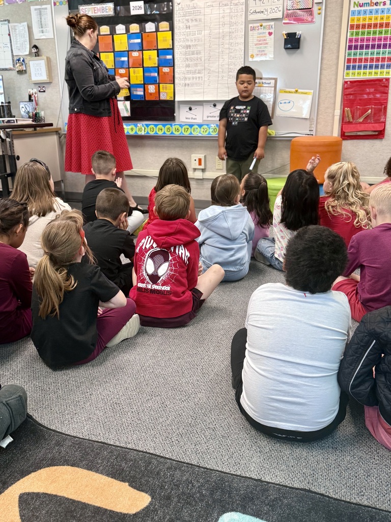 A classroom with a teacher at a whiteboard. Children sit in a circle on the floor, listening.