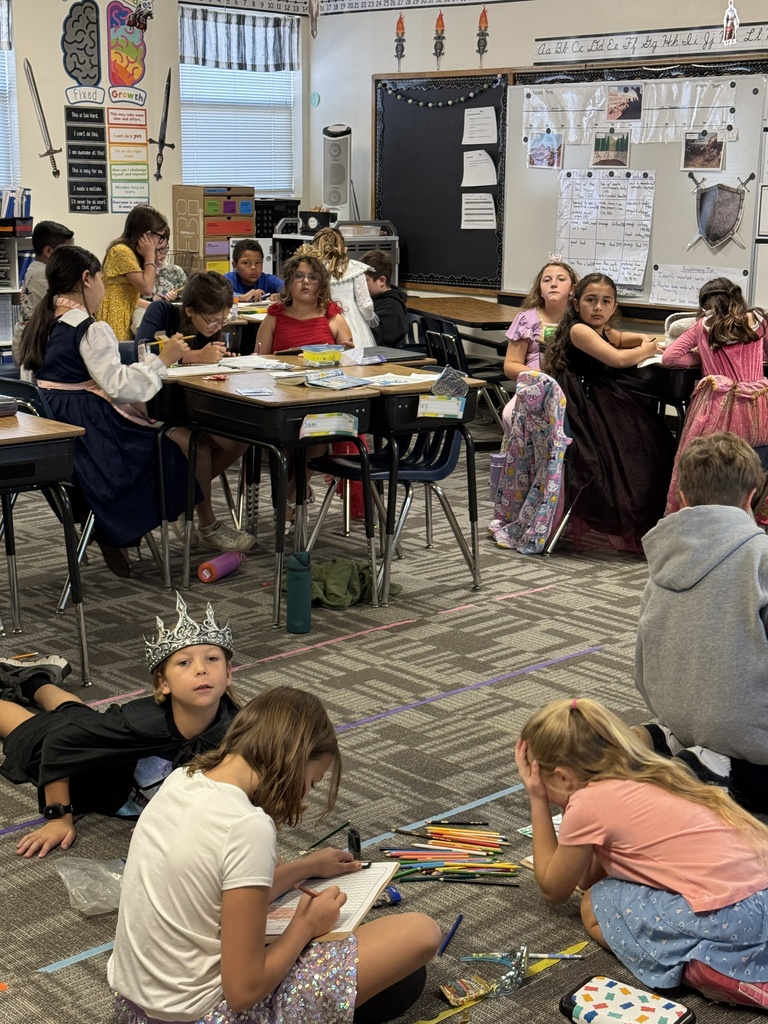 Classroom with desks and chairs; children seated and on floor. Some wear crowns. Papers, pencils, and crayons are on the floor. Bulletin boards with drawings.