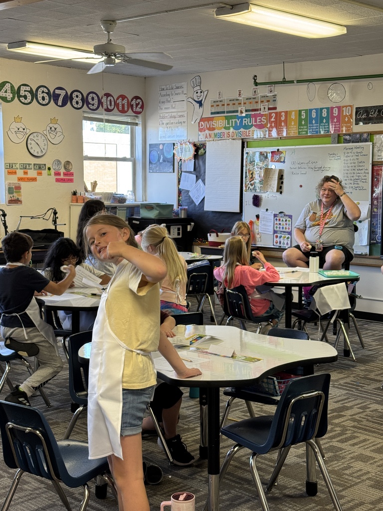 A classroom with desks and chairs; a girl stands at a desk, and others sit at tables. A teacher sits at a table, and a whiteboard with a clock is in the background.