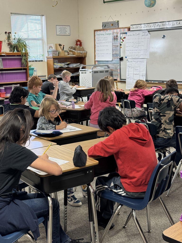 A classroom with students seated at desks. A teacher is standing at the front, near a whiteboard with writing.