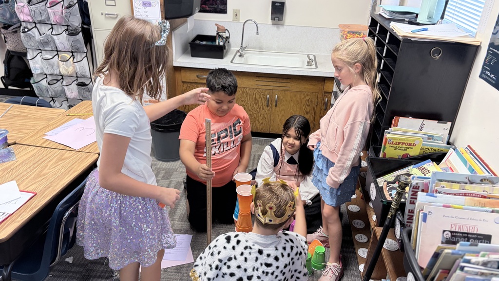 Several young children stand in a classroom. One child sits on a chair while others hold up a paper.
