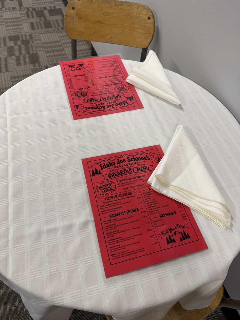 A round table with a white tablecloth, two red menus, and two white napkins. A wooden chair is on the side.