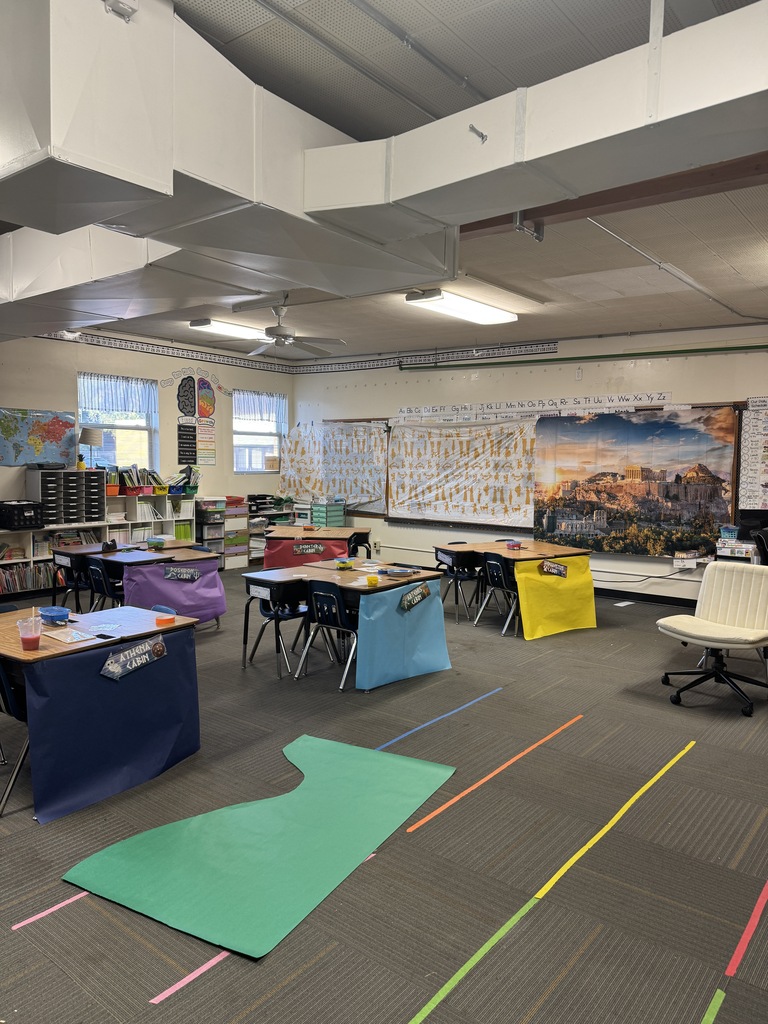 Empty classroom with desks, chairs, and tables. Colorful floor markings and maps on the wall. No people present.