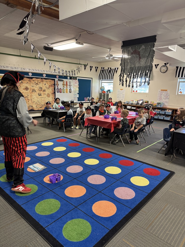 A classroom with a pirate-themed game setup. A teacher stands on a colorful game board. Students sit at tables.