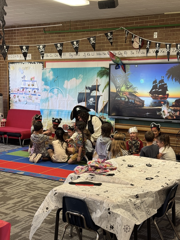 Children sit on the floor around a pirate, with a large screen behind them. A table is set up with a tablecloth.