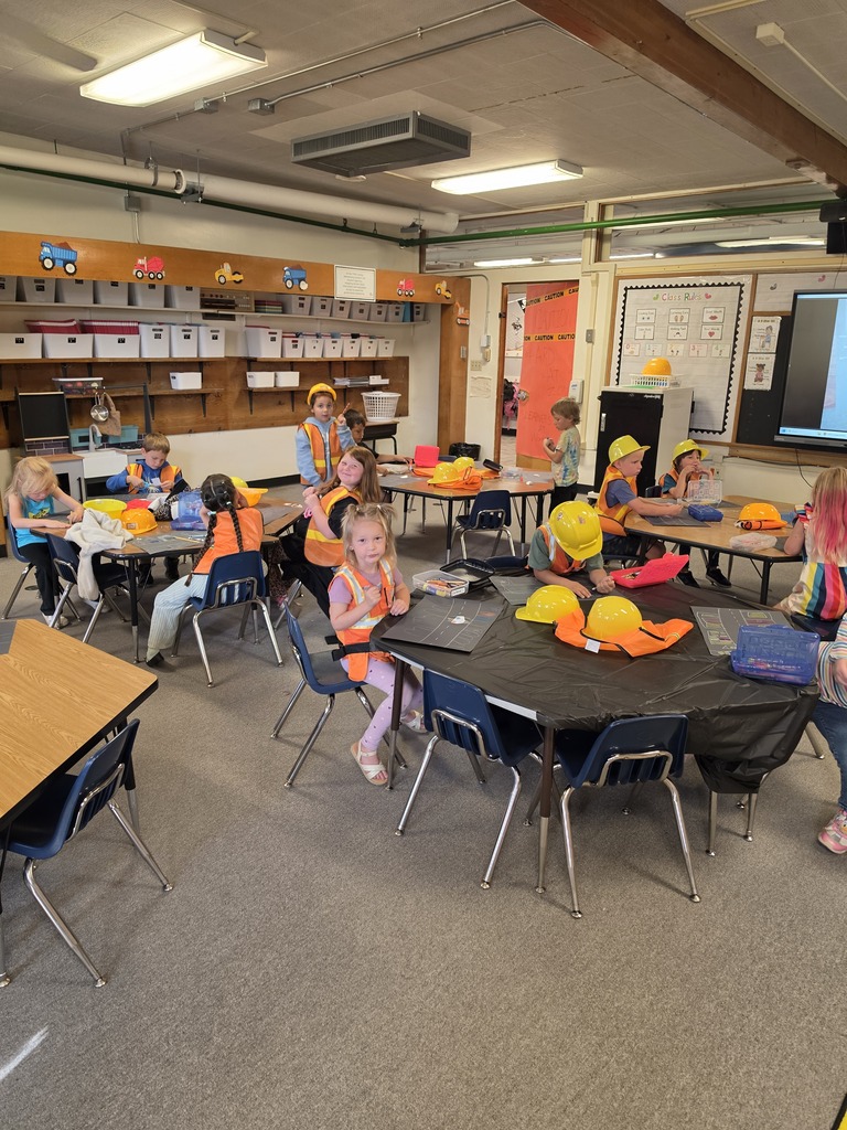 Children sit around tables in a classroom, wearing hats. The room has tables, chairs, shelves, a whiteboard, and a projector screen.