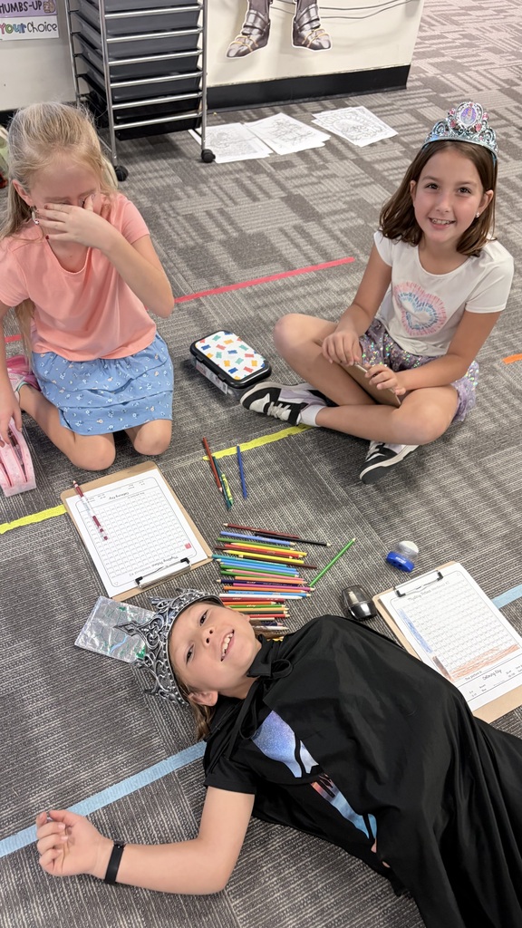 Three children in a classroom with papers and pencils on the floor. One child wears a cape, another wears a tiara.