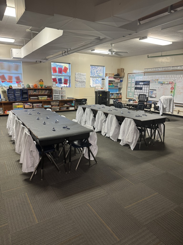 A classroom with tables and chairs, ceiling fans, and windows. Rows of white lab coats hang over tables.