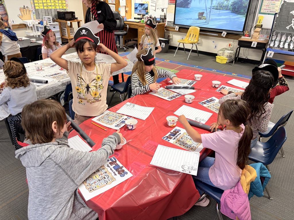 A classroom with children seated around a table with papers and supplies. Some wear hats.
