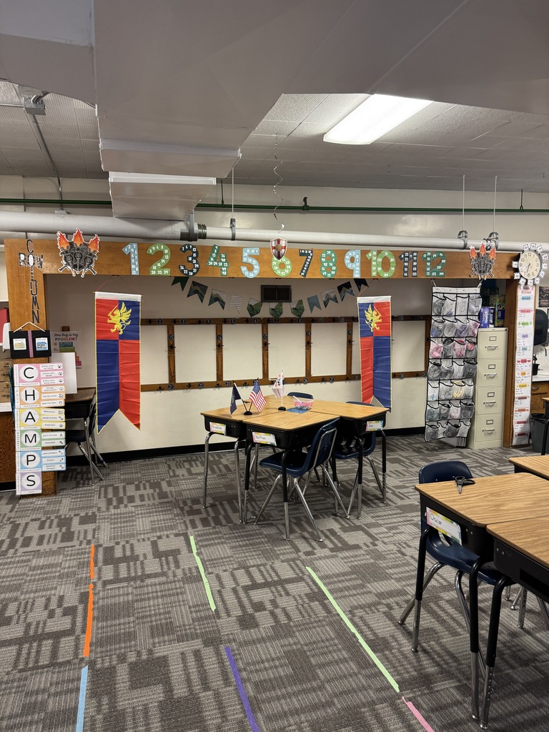 A classroom with empty tables, chairs, and a bulletin board. The ceiling has lights and decorations.