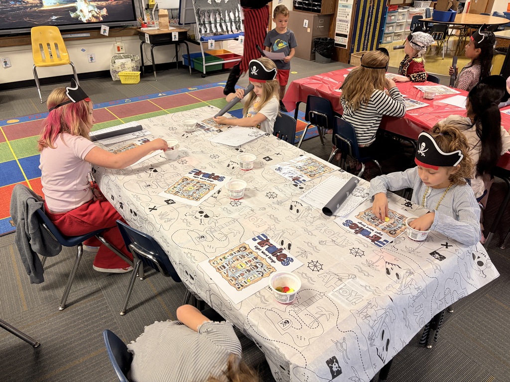 Children in a classroom wearing pirate hats, sitting at tables with maps, engaged in a group activity.